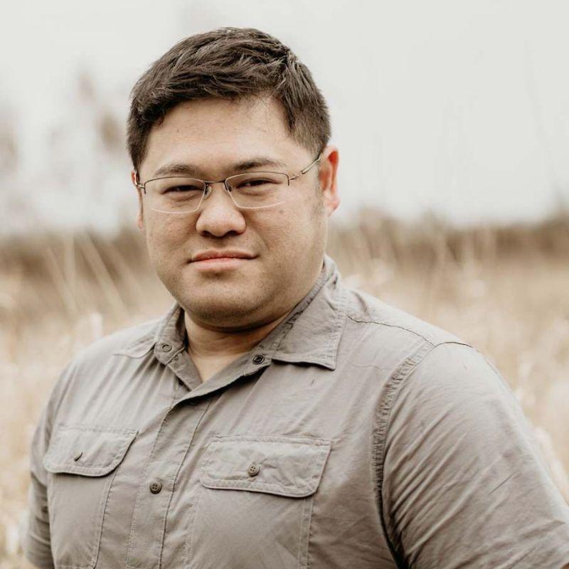 A man wearing thin-rimmed eyeglasses and a muted taupe-gray button-up shirt, photographed outdoors against a softly blurred field background.