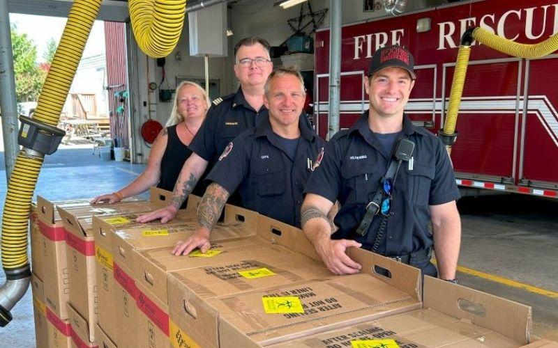 Four firefighters standing behind boxes with a fire truck in the background