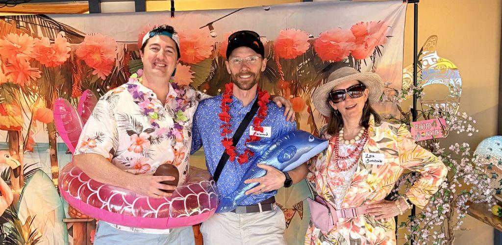 Three people posing in front of a floral backdrop with inflatable props and costumes.