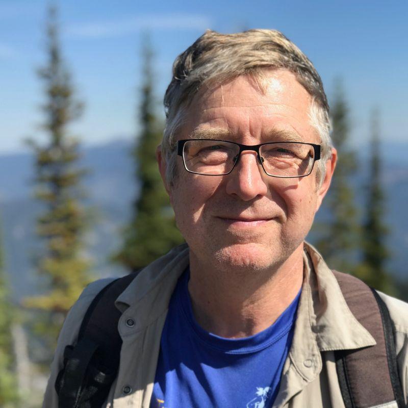 Man with glasses and a blue shirt standing outdoors with trees and mountains in the background