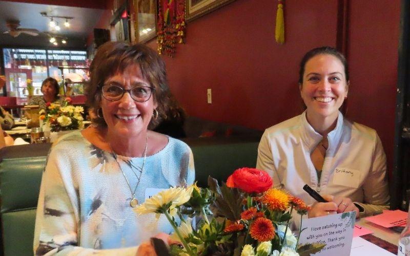 Two women sitting at a table with flowers and a card in a restaurant setting